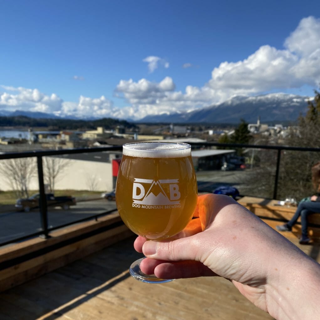 image of small glass full of beer on Dog Mountain Brewing's rooftop patio with Port Alberni in background and fluffy white clouds and snow-capped mountains contrasting blue sky