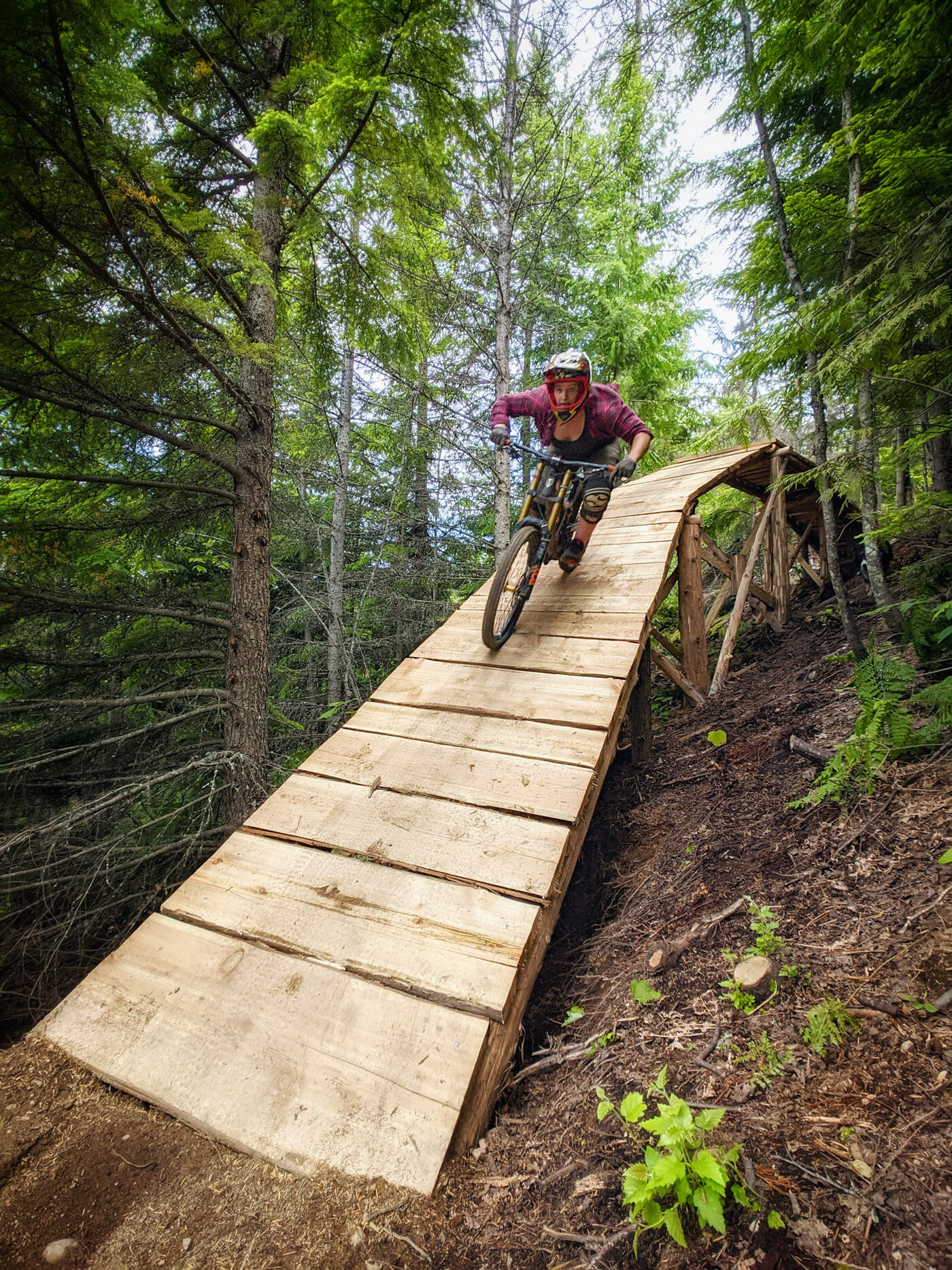 West Kootenay biking: Woman rides wooden rainbow bridge feature on forested mountain bike trail in Castlegar