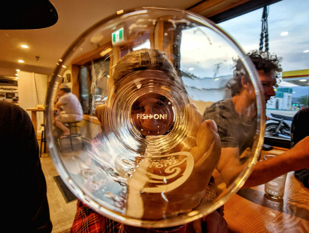 West Kootenay bikes and beer - Woman holds empty pint glass to eye with words "Fish On" on the bottom after an afternoon of mountain biking