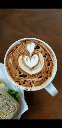 Photo of a coffee cup with coffee art in the foam, next to a sandwich in the lower left corner.