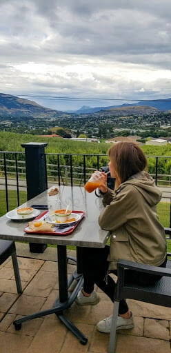 Photo of a woman sitting at a table with a food tray on it, overlooking the Okanagan Valley as she sips on a drink.