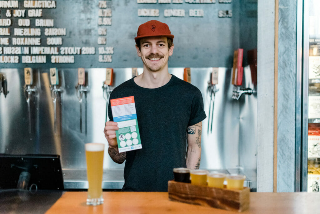 A staff member showing a stamped passport along with a flight and pint at Strange Fellows Brewing